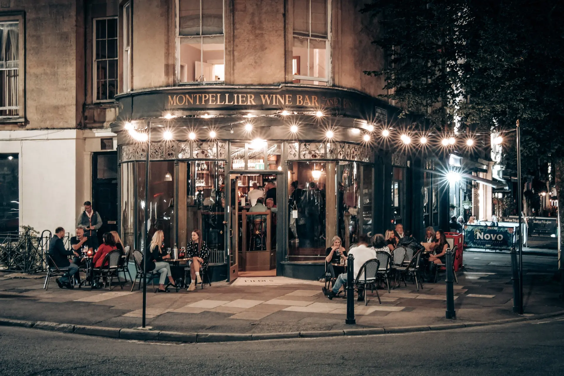 The Montpellier Wine Bar at night, festoon lights strung above pavement tables, with the apartment windows lit above